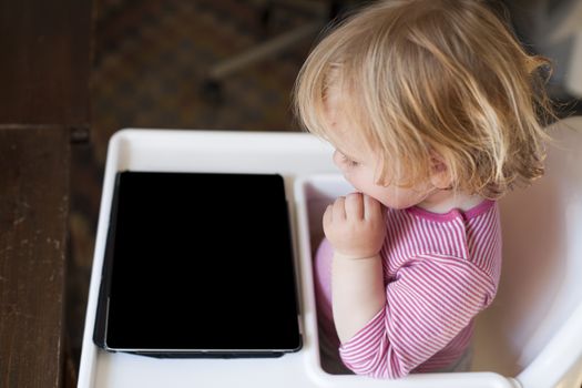 blonde nineteen month age baby red and white striped shirt looking at blank black screen digital tablet sitting in high chair inside home