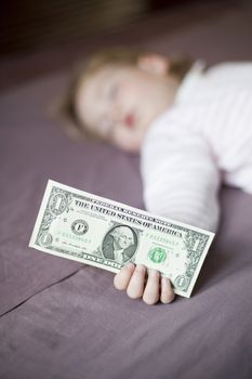 closeup one dollar banknote in hand of blonde caucasian baby nineteen month age with pink and white stripped jersey sleeping on brown sheets king bed
