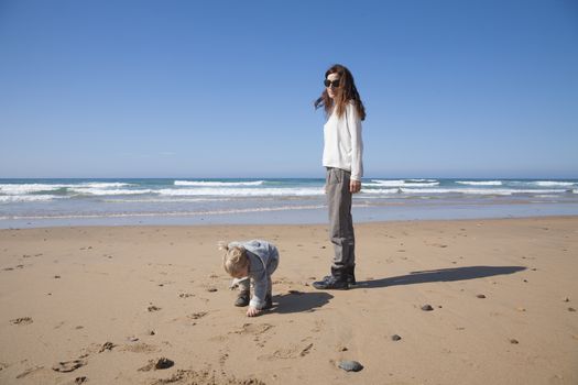 two years age blonde baby with grey coat and brunette mother woman walking in sand of beach in front of water sea or ocean