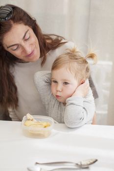 portrait of two years age blonde baby with grey shirt eating chicken from tupperware sitting on legs of mother woman in white table