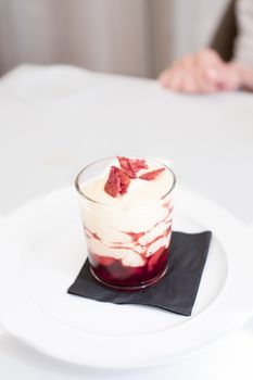 still life of delicious sweet haute cuisine dessert with white cream strawberry sauce jam and red cracker inside glass over black paper napkin and white dish on restaurant white table