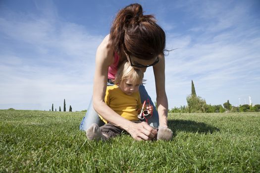 brunette woman mother tying shoelaces to two years aged blonde baby with yellow shirt with white kid sunglasses sitting on green grass lawn in park