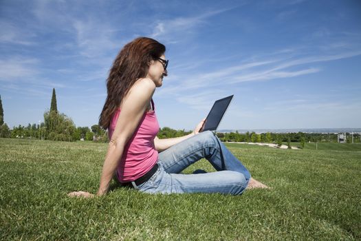redhead woman blue jeans trousers pink shirt reading digital tablet blank screen sitting on green grass lawn in park