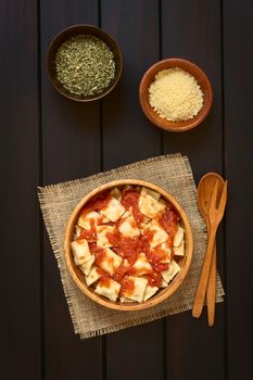 Cooked ravioli with homemade tomato sauce in wooden bowl with grated cheese and dried oregano in small bowls, wooden spoon and fork on the side, photographed overhead on dark wood with natural light