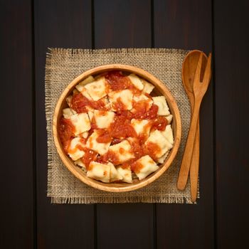 Cooked ravioli with homemade tomato sauce in wooden bowl with wooden spoon and fork on the side, photographed overhead on dark wood with natural light