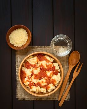 Cooked ravioli with homemade tomato sauce in wooden bowl with grated cheese and glass of water, wooden spoon and fork on the side, photographed overhead on dark wood with natural light