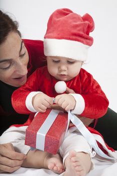 one year age caucasian blonde baby Santa Claus disguise with brunette woman mother red cardigan green trousers opening Christmas box gift on white background