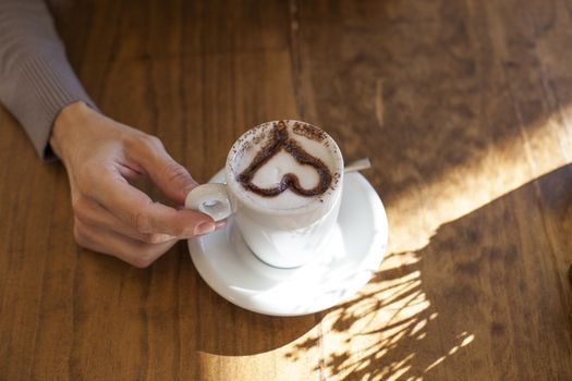 white small cup cappuccino coffee with chocolate heart in woman hands on light brown wooden table