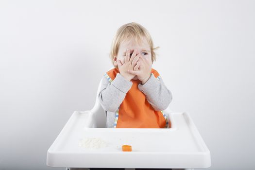 blonde caucasian baby seventeen month age orange bib grey sweater eating meal in white high-chair with hands on eyes does not like carrot