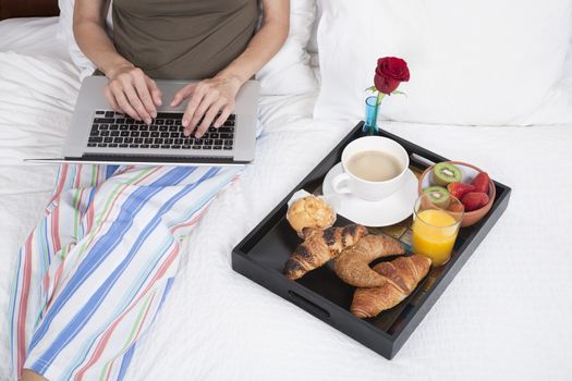 woman green shirt striped pajama pants sitting on white bed typing keyboard laptop with breakfast tray croissants orange juice strawberry kiwi cupcake red rose flower