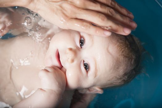 six months age blonde baby body and face washing by woman mother hands in blue little plastic bath indoor with brown background