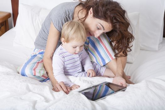 eighteen month aged blonde baby with brunette woman mother reading digital tablet on white bed
