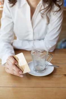 woman with white shirt ready to pay empty finished cappuccino coffee cup with made up credit card in hand on light brown wooden table cafe