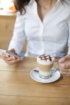 woman touching mobile phone with cappuccino coffee cup on light brown wooden table cafe