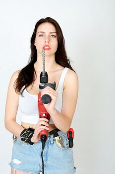 Young female model wearing white top and jeans' shorts. Girl holding big driller, keeping key, screwdriver and small hammer in her pocket.