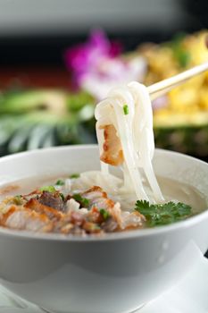 Closeup of a person eating Thai style crispy pork rice noodle soup from a bowl with chopsticks. Pineapple fried rice in the background.