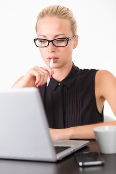 Business woman wearing black glasses, holding a pen in her hand, checking some information on her laptop computer in office. 