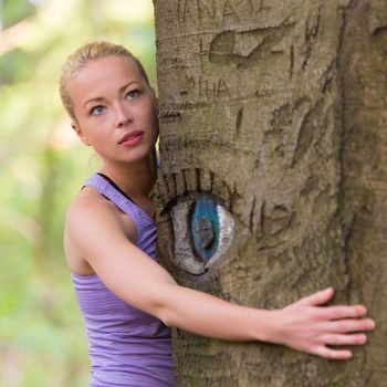 Relaxed young lady embracing a tree receiving life energy from the nature. Eye carved in tree trunk.