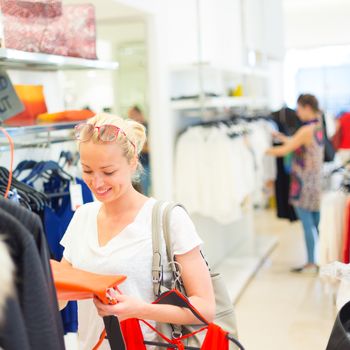 Woman shopping clothes. Shopper looking at clothing indoors in store. Beautiful blonde caucasian female model wearing casual clothes and fashionable sunglasses. Focus on model, shallow depth of field.