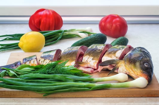 On the table on the kitchen blackboard is cleaned and prepared for cooking fish. Nearby are the components for its preparation: cucumbers, onions, dill, tomatoes, lemon.