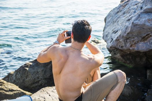 Rear View of a Shirtless Young Man Taking Photos at the Beach While Sitting on Big Rock Under the Heat of the Sun.