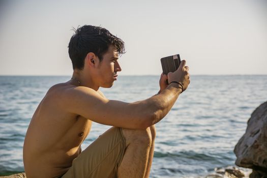 Side View of a Shirtless Young Man Taking Photos at the Beach While Sitting on Big Rock Under the Heat of the Sun.