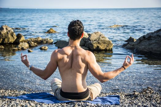 Handsome Shirtless Young Man During Meditation or Doing an Outdoor Yoga Exercise Sitting on Rocks by Ocean Shore