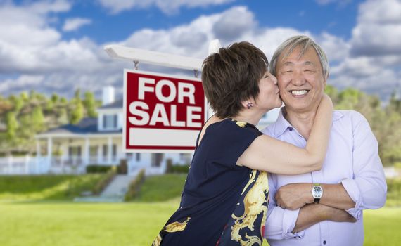 Attractive Affectionate Senior Chinese Couple In Front of Beautiful House and For Sale Real Estate Sign.