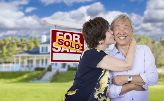 Attractive Affectionate Senior Chinese Couple In Front of Beautiful House and Sold For Sale Real Estate Sign.