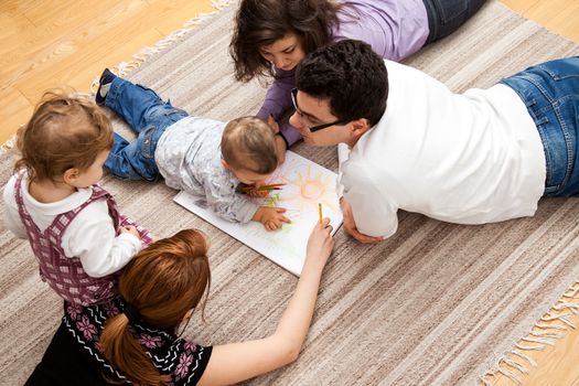 family group of five - two babies and three adults lying on the carpet, drawing a picture together.