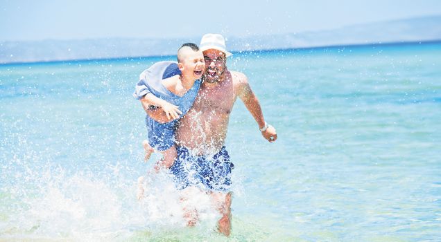 Father holding his son is running through sea or ocean, splashing water arround.