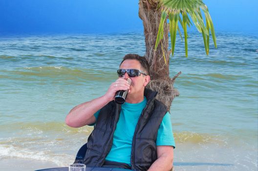 A man wearing sunglasses sitting in front of a palm tree. In the background the sea.