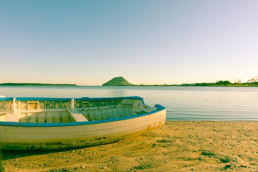 Old faded effect image clinker dinghy catching morning sun on beach with Mount Maunganui on horizon.