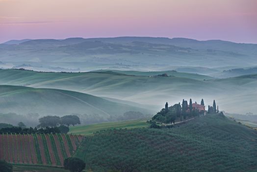 Foggy sunrise with a pink sky and mist between the Tuscan hills over agriturismo podere il Belvedere farmhouse in the Valdorcia (Orcia Valley) between Pienza and San Quirico in Tuscany, Italy.