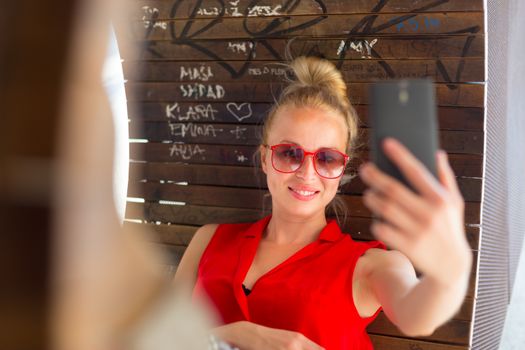 Casually dressed young cheerful lady wearing red sunglasses taking carefree selfie portrait outdoors with her android smarthphone to post it on social networks.