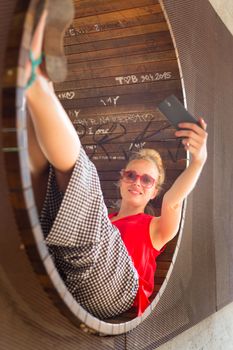 Casually dressed young cheerful lady wearing red sunglasses taking carefree selfie portrait outdoors with her android smarthphone to post it on social networks.