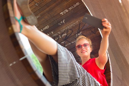 Casually dressed young cheerful lady wearing red sunglasses taking carefree selfie portrait outdoors with her android smarthphone to post it on social networks.