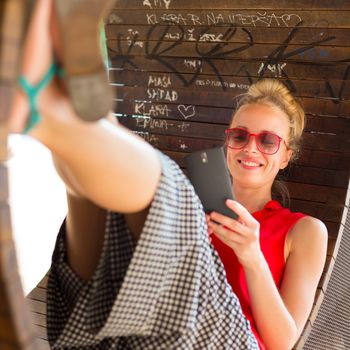 Casually dressed young cheerful lady wearing red sunglasses playing carelessly with her android smarthphone browsing trough social networks relaxing on  contemporary circular bench in city park.