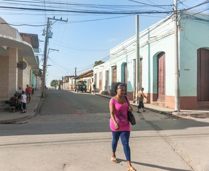 Trinidad, Cuba - July 2, 2012: Woman in pink top stands out as she walks along street lined by colonial style in Cuban town where others are sitting on corner