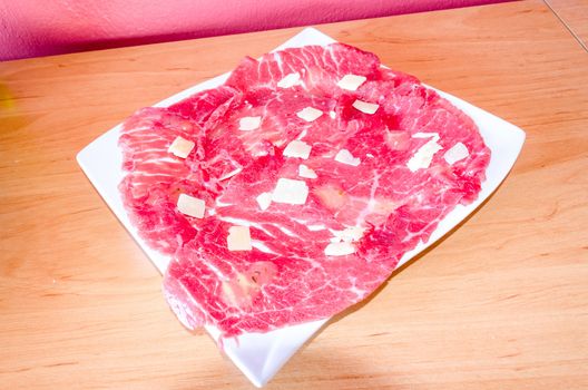 White dish with carpaccio of beef with parmesan. On a wooden base.
