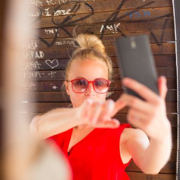 Casually dressed young cheerful lady wearing red sunglasses taking carefree selfie portrait outdoors with her android smarthphone to post it on social networks.