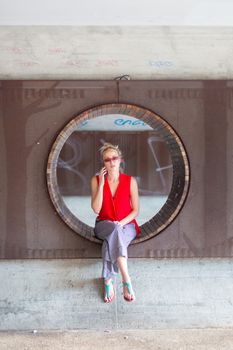 Casually dressed young cheerful lady wearing red sunglasses talking on mobile phone sitting on contemporary circular bench in city park.