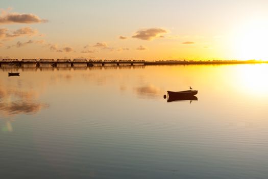 Sunrise and calmness over Tauranga Harbour. Water ripples bird on boat and historic rail bridge on horizon.