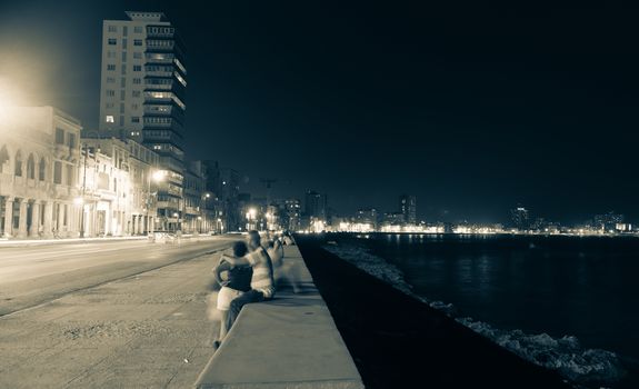 Along the Malecon, Havana, Cuba, people out at night in warm sea air sitting on sea wall across from the colonial buildings with the more modern city lights in distance.