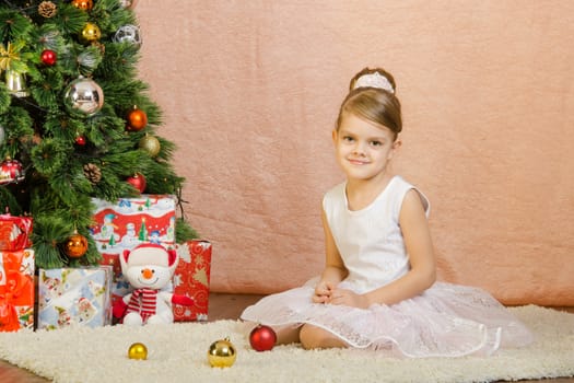Five-year girl in a New Year's dress sitting on a mat at the Christmas tree