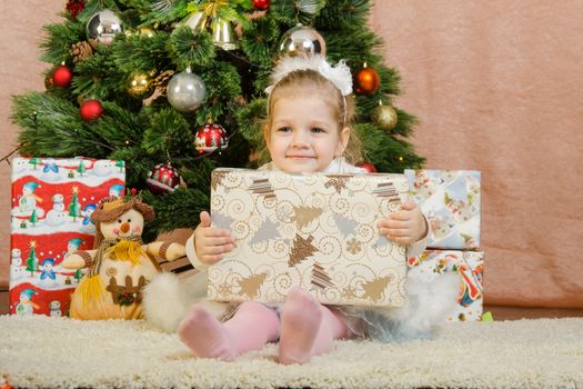 The three-year girl in a New Year's dress sitting on a mat at the Christmas tree