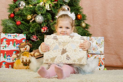 The three-year girl in a New Year's dress sitting on a mat at the Christmas tree