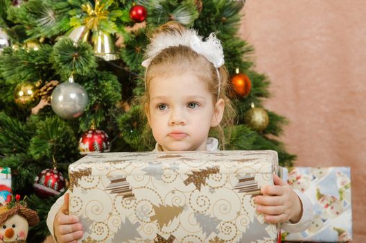 The three-year girl in a New Year's dress sitting on a mat at the Christmas tree