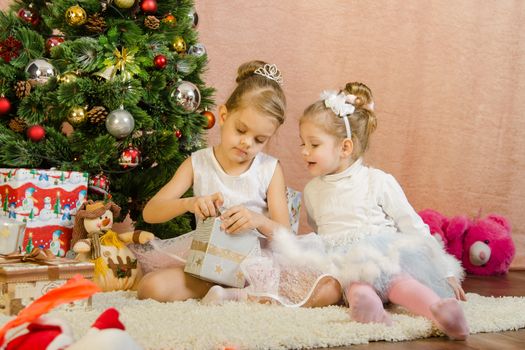 Five-year and three-year girl in a New Year's dress sitting on a mat at the Christmas tree