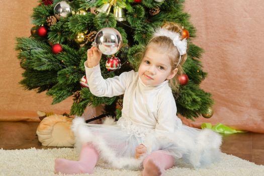 The three-year girl in a New Year's dress sitting on a mat at the Christmas tree